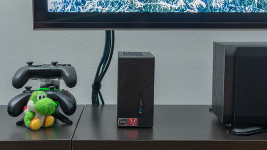 A small computer standing on a TV bench next to a pair of game controllers and a speaker.